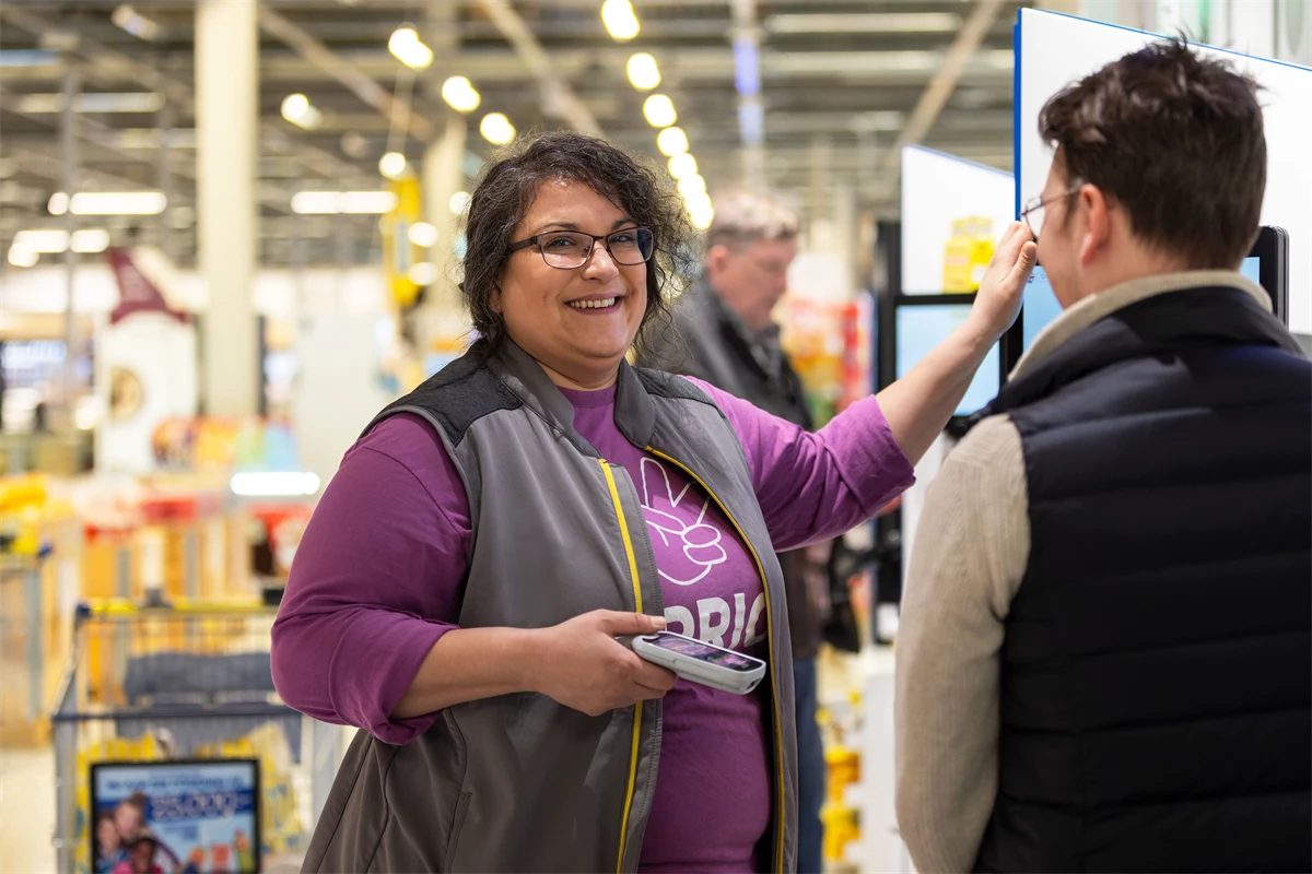 En kvinna som jobbar i matbutik, hjälper en kund vid självskanningskassorna.
