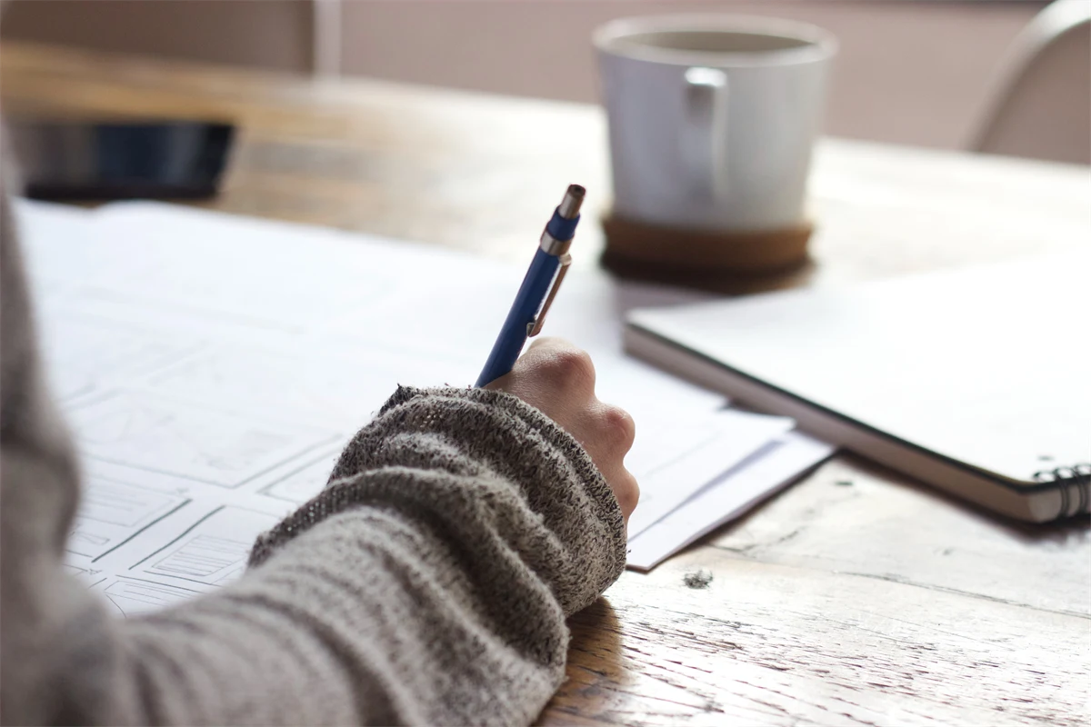Närbild av ett skrivbord med kaffekopp, anteckningsblock och en kvinnas hand som skriver med en penna på papper