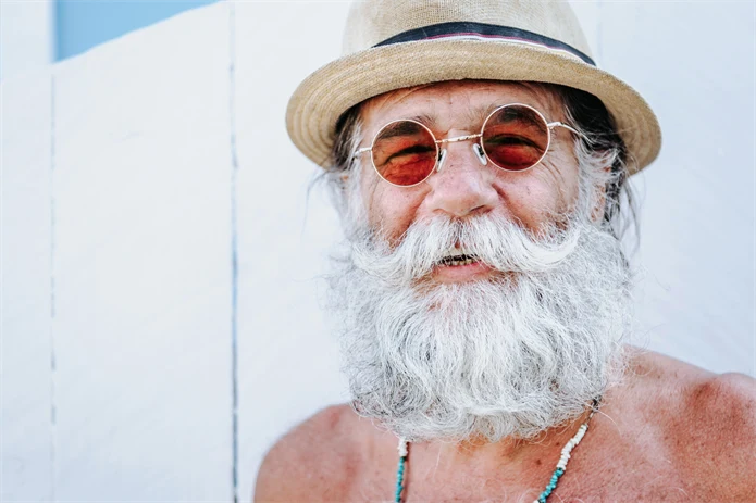 Glad äldre man med lekfullt vitt skägg, runda solglasögon och stråhatt på stranden.