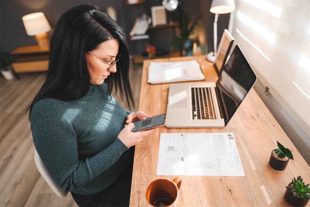 Kvinna sitter vid skrivbord med dator och mobiltelefon.